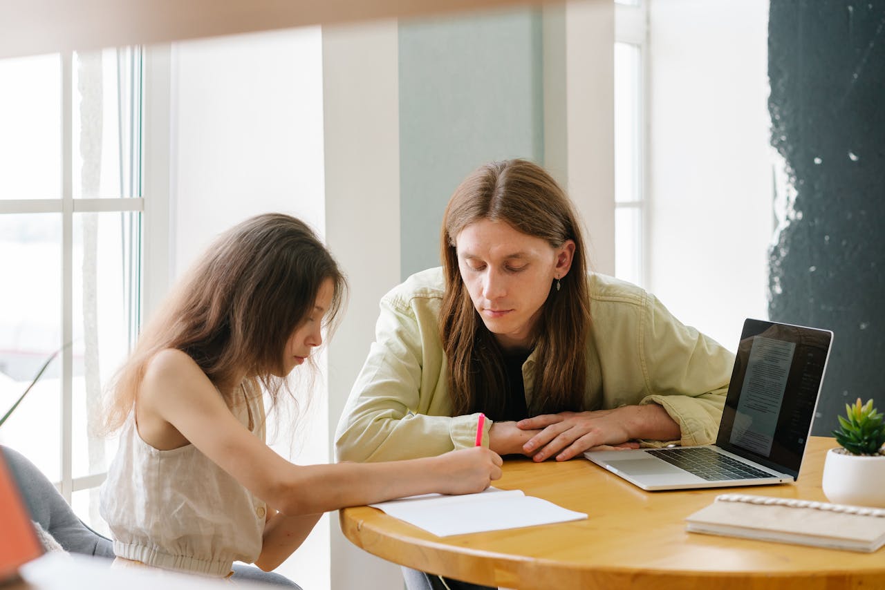 services-01 A father helps his daughter with homeschooling indoors, featuring a laptop and study materials.
